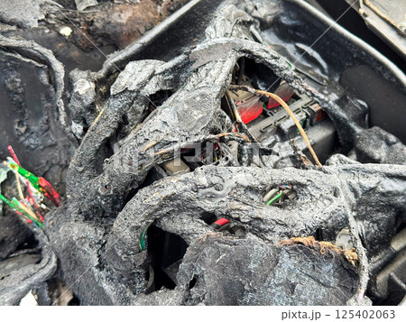 Burned car fuse box and wiring with melted insulation, scorched plastic housing, and exposed colored wires after extensive fire damage in the engine compartment 125402063