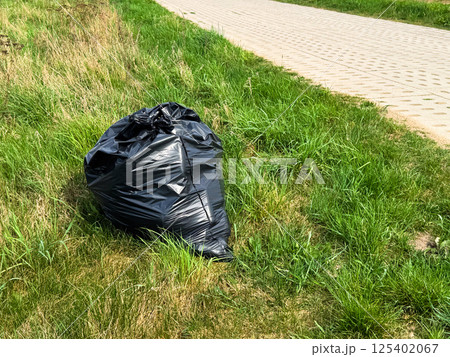 Large black garbage bag placed on green grass near a paved sidewalk, representing outdoor litter or cleanup effort in a park or urban green space during spring 125402067