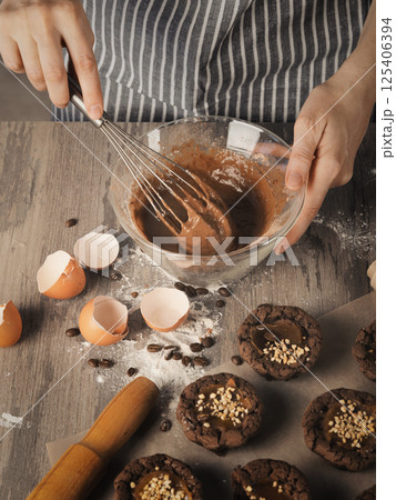 Female hands beat with a whisk dough into a bowl next to muffins, eggshell and rolling pin on on a table, faceless baker in striped apron, vertical image. 125406394