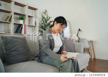 Asian young woman with glasses reading a book on the sofa in the living room happily Asian young woman with glasses reading a book on the sofa in the living room happily 125406929