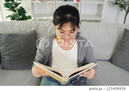 Asian young woman with glasses reading a book on the sofa in the living room happily Asian young woman with glasses reading a book on the sofa in the living room happily 125406930