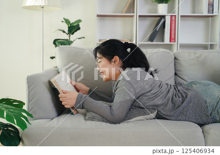 Asian young woman with glasses reading a book on the sofa in the living room happily Asian young woman with glasses reading a book on the sofa in the living room happily 125406934