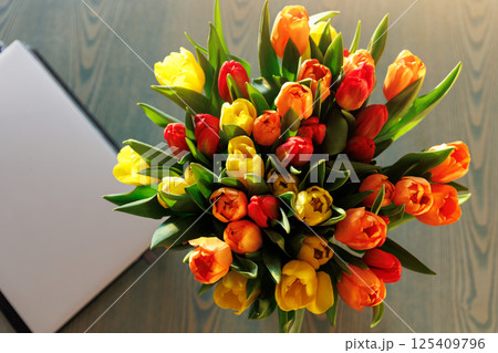 Top above view fresh multicolored tulips bouquet  in vase table closed laptop warm sunset sun lights against balcony window cozy home interior. Blooming flowers decoration  living room provence style 125409796