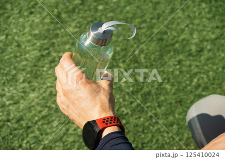 Sport man holding water bottle on outdoor sports ground with green grass. Concept water balance for heat season for workout. Close up. Copy space. 125410024
