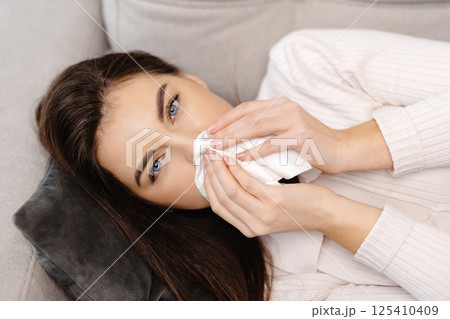Unhappy sad young female with plaid, suffering from fever and flu on sofa, blowing nose in napkin in living room interior Unhappy sad young female with plaid, suffering from fever and flu on sofa, blowing nose in napkin in living room interior 125410409