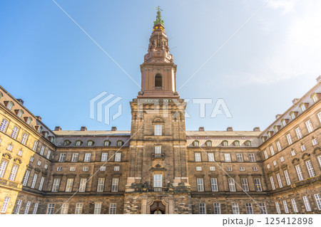 Visitors admire the stunning architecture of Christiansborg Palace in Copenhagen, surrounded by historical charm under a bright blue sky. The grandeur of this government building captivates all. 125412898