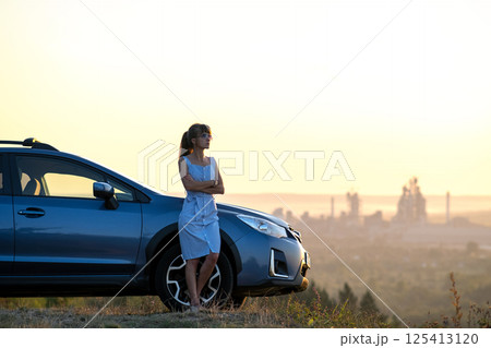 Happy young woman in blue dress standing near her vehicle looking at sunset view of summer nature. Travelling and vacation concept. 125413120