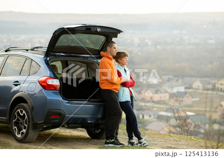 Happy couple standing together near a car with open trunk enjoying view of rural landscape nature. Man and woman leaning on family vehicle luggage compartment. Weekend travel and holidays concept. Happy couple standing together near a car with open trunk enjoying view of rural landscape nature. Man and woman leaning on family vehicle luggage compartment. Weekend travel and holidays concept. 125413136