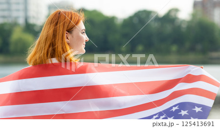 Back view of happy young woman with USA national flag on her shoulders. Positive girl celebrating United States independence day. 125413691