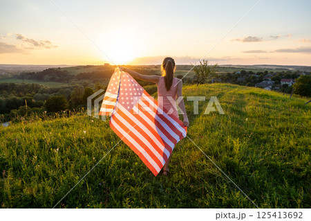 Back view of happy woman with USA national flag standing outdoors at sunset. Positive female celebrating United States independence day. International day of democracy concept. Back view of happy woman with USA national flag standing outdoors at sunset. Positive female celebrating United States independence day. International day of democracy concept. 125413692