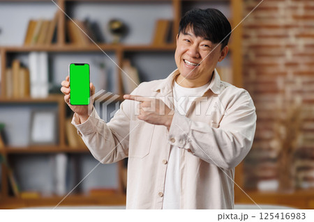 happy asian man, showing device, Online offer, empty free space, cheerful man with short black hair stands in a warm, inviting room, pointing at a mobile device that features a green screen, inviting 125416983