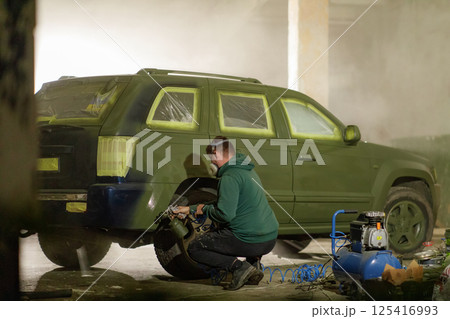 A person is spray painting an SUV in a dim garage. Most of the vehicle is covered in a protective layer, while the individual focuses on detailing the tire area with precision. A person is spray painting an SUV in a dim garage. Most of the vehicle is covered in a protective layer, while the individual focuses on detailing the tire area with precision. 125416993