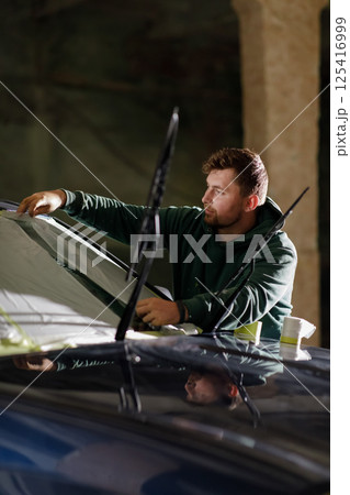 A man focuses on replacing the windshield of a car in a dimly lit area during nighttime. He is carefully adjusting the glass and ensuring a proper fit. A man focuses on replacing the windshield of a car in a dimly lit area during nighttime. He is carefully adjusting the glass and ensuring a proper fit. 125416999