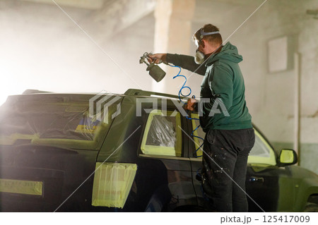 A person is working on repainting a car in a dimly lit workshop. They are wearing a mask and goggles while spraying paint, ensuring proper coverage and safety precautions. 125417009