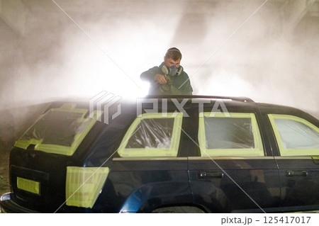 A man is applying paint to a black SUV in a workshop, using spray equipment while the car is masked with tape and plastic. The environment is misty, enhancing the focus on his work. A man is applying paint to a black SUV in a workshop, using spray equipment while the car is masked with tape and plastic. The environment is misty, enhancing the focus on his work. 125417017