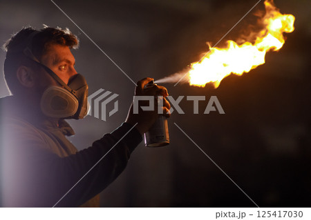 A person in a protective mask is igniting a spray can, producing a bright flame in a low-light workshop. Sparks fly as he focuses on his task carefully. A person in a protective mask is igniting a spray can, producing a bright flame in a low-light workshop. Sparks fly as he focuses on his task carefully. 125417030