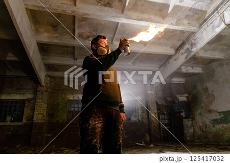 A person in a dark, abandoned industrial space uses a spray can to produce a flame. The dim surroundings highlight the fire's glow, showcasing the contrast between light and shadow. 125417032
