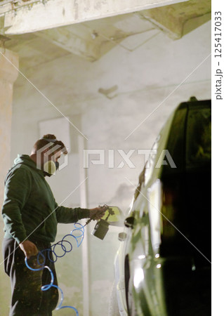 A person wearing a protective mask is carefully spraying a car with a handheld device in a dimly lit garage, focusing on achieving a smooth finish. The atmosphere is filled with spray mist. A person wearing a protective mask is carefully spraying a car with a handheld device in a dimly lit garage, focusing on achieving a smooth finish. The atmosphere is filled with spray mist. 125417033