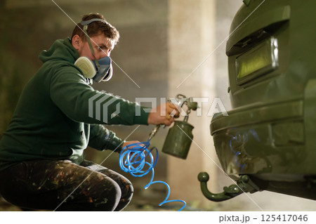 A person is carefully applying paint to a car using a spray gun while wearing a mask for protection. The workshop is dimly lit, highlighting the meticulous details of the process. A person is carefully applying paint to a car using a spray gun while wearing a mask for protection. The workshop is dimly lit, highlighting the meticulous details of the process. 125417046