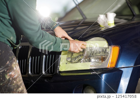 A person is carefully applying tape around a car headlight in a garage, focused on restoring its shine. The workspace is well-lit, highlighting the effort in this maintenance task. A person is carefully applying tape around a car headlight in a garage, focused on restoring its shine. The workspace is well-lit, highlighting the effort in this maintenance task. 125417058