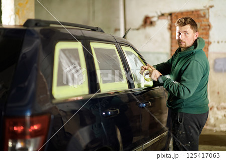 A man applies masking tape to the windows of a vehicle in a workshop setting during the evening. He is intent on ensuring a precise paint job as he works methodically. 125417063