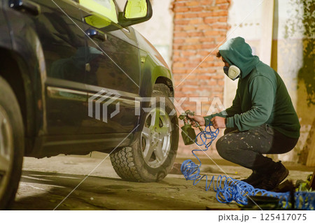 A person in a green hoodie and mask is crouching beside a vehicle, using an air tool to inflate a tire. The surroundings are a garage with exposed brick and concrete, suggesting a working atmosphere. 125417075