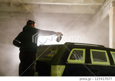 A person is spray painting a car in a dimly lit industrial workshop at night. The misty air and protective coverings create an artistic atmosphere while the vehicle is being prepped for a fresh coat. 125417077