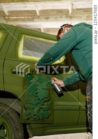 An artist is focused on spray painting a vintage car using a camouflage stencil. The setting showcases an urban environment with brick walls and scattered art supplies. 125417085