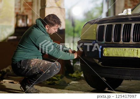 A man in a green hoodie kneels beside a black Jeep in a dimly lit industrial space, carefully preparing the front bumper for detailing as evening light filters in. A man in a green hoodie kneels beside a black Jeep in a dimly lit industrial space, carefully preparing the front bumper for detailing as evening light filters in. 125417097