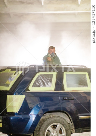 An individual wearing a mask stands on the roof of a vehicle, preparing to apply spray paint. The garage is filled with smoke, creating a dramatic atmosphere for the project. 125417110