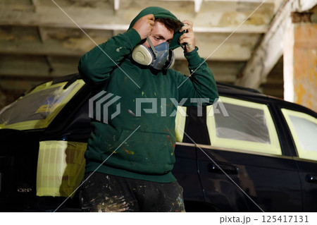 In an industrial garage, a worker puts on a mask while standing near a black car that's being painted. The area shows evidence of paintwork and preparation for the task ahead. In an industrial garage, a worker puts on a mask while standing near a black car that's being painted. The area shows evidence of paintwork and preparation for the task ahead. 125417131