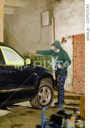 A person is carefully painting a dark vehicle in a garage. Wearing a mask and hoodie, the individual uses a spray gun to apply paint, ensuring precision on the car's surface. 125417143