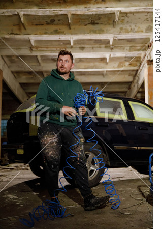 A man stands in a dusty garage, holding a coiled blue air hose and wearing a hooded sweatshirt, surrounded by the parts of a vehicle being restored. The atmosphere is industrial and focused. 125417144