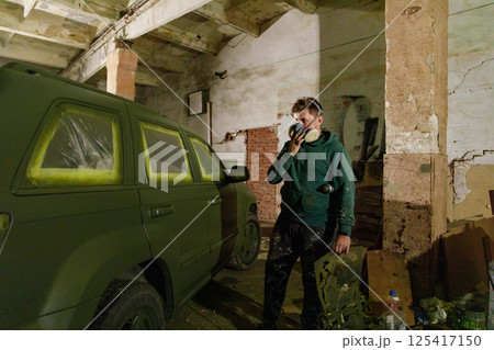A worker is wearing a mask while carefully painting a large vehicle with camouflage colors in a dim, industrial space illuminated by warm light. Cardboard pieces and tools are nearby. 125417150