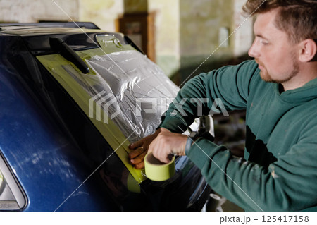 Individual is applying protective plastic and tape to a vehicle window in a dimly lit garage. The activity is focused and methodical as the person works carefully on the task. 125417158