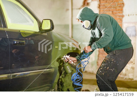 A masked individual meticulously sprays paint onto a vehicle's surface in a workshop. The low lighting adds a rugged atmosphere as focus is on the task of restoration and refinishing. 125417159
