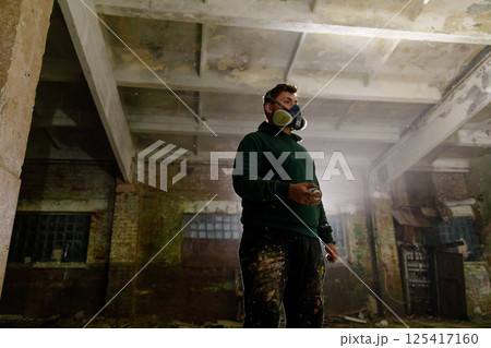 An urban artist stands in an abandoned warehouse, using spray paint to create artwork. Wearing a protective mask, the artist is surrounded by dust and debris, showcasing their creative process. 125417160