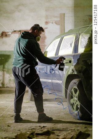 A person wearing a mask spray paints a car inside an old workshop. The environment is dusty and industrial, highlighting the focus and effort in car customization. 125417188