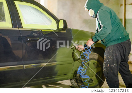 A person in a hoodie applies paint to a car using a spray gun in a garage setting. The focus is on the meticulous work being done to rejuvenate the vehicle's appearance after thorough preparation. 125417218