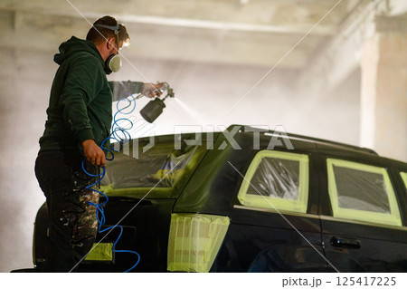 A man is spray painting a vehicle inside a dimly lit workshop. He carefully uses a spray gun, while sections of the car are masked off with tape for precise detailing. A man is spray painting a vehicle inside a dimly lit workshop. He carefully uses a spray gun, while sections of the car are masked off with tape for precise detailing. 125417225