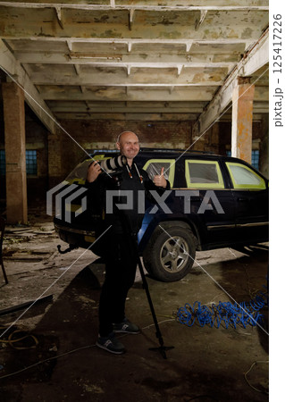 In a dimly lit, deserted industrial space, a photographer strikes a playful pose with his camera beside a parked black SUV, surrounded by rustic elements and scattered debris. 125417226