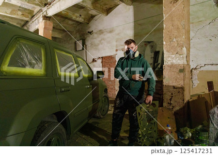 In a shadowy garage, a man wearing a gas mask applies green paint to an SUV, focused on transforming its appearance with a vibrant design in a hands-on project. 125417231