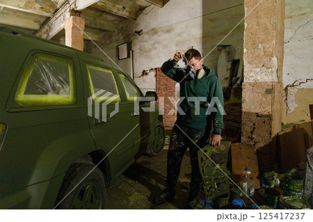 A person wearing a gas mask stands next to a partially painted green vehicle inside a warehouse. The ambient lighting and surroundings convey an atmosphere of urban artistry and transformation. 125417237