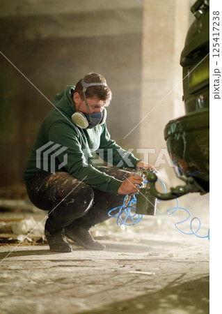 A skilled worker is crouched on the floor, meticulously painting a vehicle with a spray gun. The workshop is filled with dust and soft light highlighting the process of automotive refinishing. 125417238