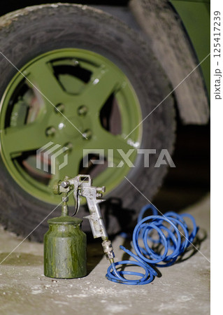 A spray application tool rests beside a green canister, with a blue air hose coiled on the ground next to a vehicle's green wheel in a dimly lit area. 125417239
