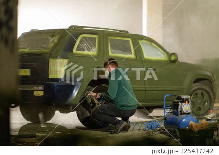 A man concentrates on painting a green SUV in a workshop. The surroundings are dimly lit with tools and paint visible, illustrating an engaging restoration process during the evening. 125417242