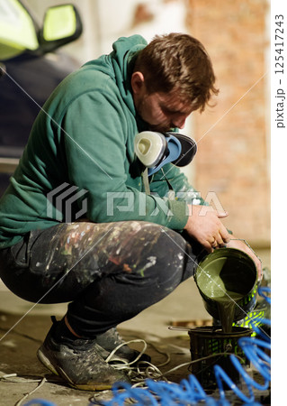 Focused artist in a green hoodie mixes paint in a rustic workshop. The evening light hints at a creative mural project nearing its start in an industrial space. Focused artist in a green hoodie mixes paint in a rustic workshop. The evening light hints at a creative mural project nearing its start in an industrial space. 125417243