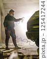 A person in a mask paints a car with spray paint in a dilapidated warehouse, surrounded by debris and a soft glow of light illuminating the scene, highlighting a moment of artistic expression. 125417244