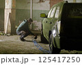A worker is carefully painting a vehicle inside a dimly lit, abandoned warehouse. The setting features rough walls and scattered debris, highlighting the restoration effort underway. 125417250