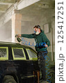 A man in a green hoodie wears a mask and goggles while using a spray gun to paint a vehicle inside a workshop. Protective tape covers parts of the car for precision work. 125417251